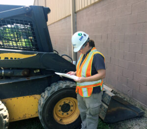 Stephanie standing next to a piece of equipment, looking at a clipboard, wearing her hard hat & safety vest.