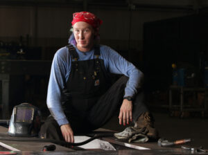 Melissa poses for a portrait wearing a red bandana, blue long sleeve shirt, and black overalls. She is sitting & has pink painted fingernails.