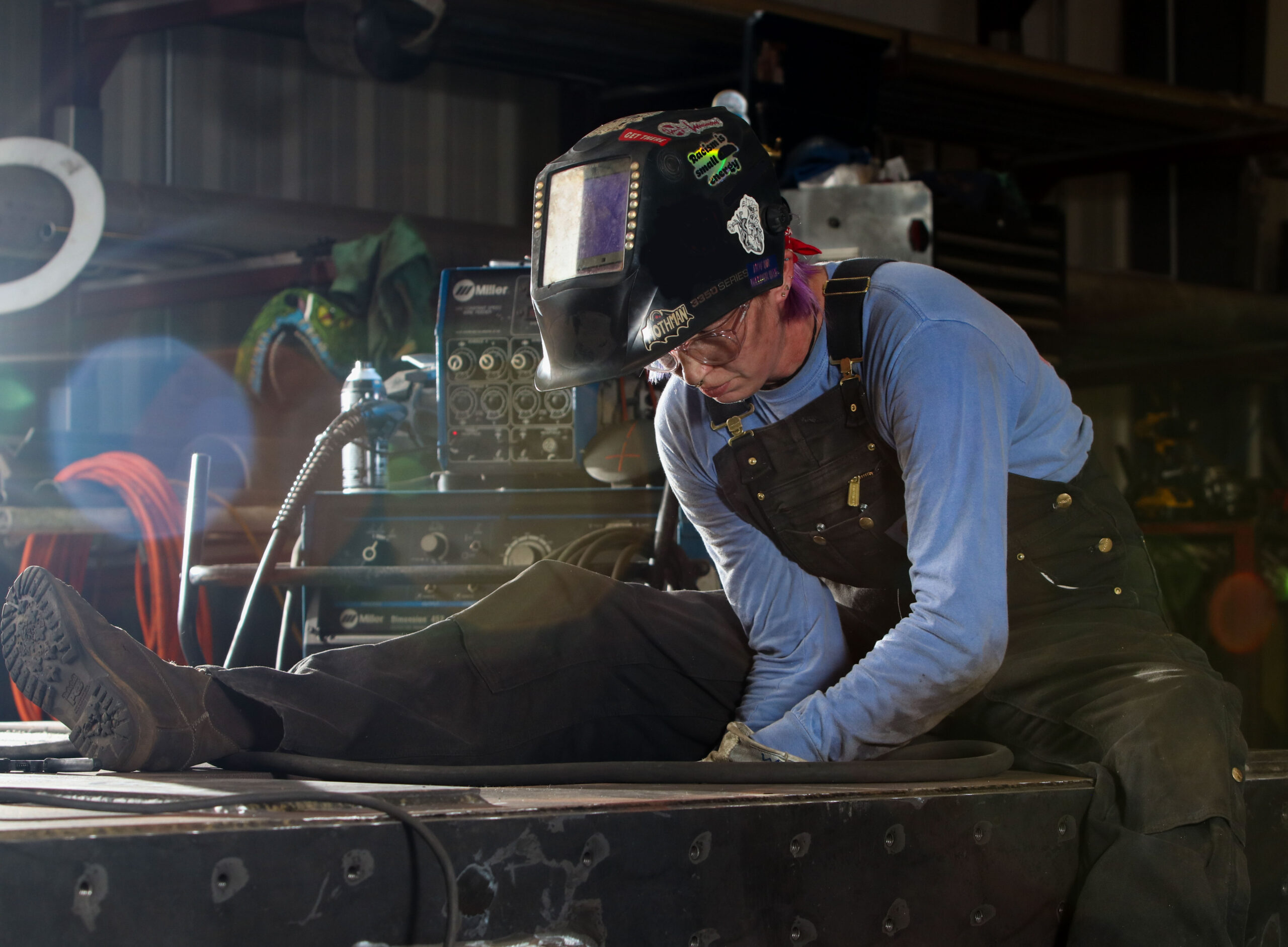 Melissa wearing a welding hood, long sleeve blue shirt, and dark overalls. She is mimicking welding for the photo.
