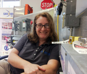 Stephanie looking at the camera & smiling. Wearing a blue shirt & leaning against the LOTO board.